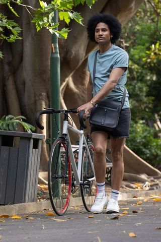 Urban cyclist walking bike through leafy park wearing teal shirt and crossbody bag