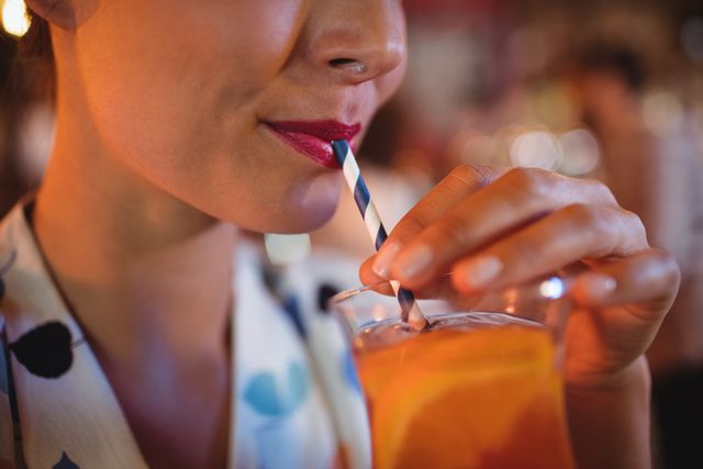 This image captures a close-up of a woman sipping a cocktail through a striped straw in a pub. Her red lipstick and the vibrant colors of the drink add a lively touch. Ideal for use in advertisements for bars, nightlife promotions, social media posts about leisure activities, or articles on cocktail recipes and summer drinks.