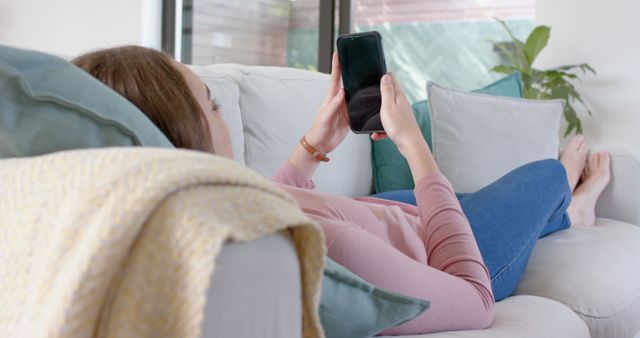 A woman is lying on a sofa in a cozy home setting, using a smartphone. This scene can be used to depict relaxation, modern technology usage, lifestyle, and home comfort. Ideal for advertisements or editorial content about home living, mobile technology, and daily life.