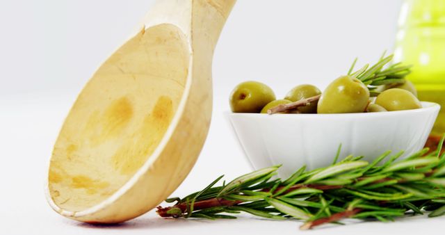 Close-up of a wooden spoon resting near a white bowl filled with green olives and fresh rosemary sprigs. Ideal for use in cooking blogs, healthy eating websites, Mediterranean diet promotions, and culinary magazines.