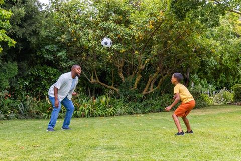 African american father coaching youth son playing soccer on garden lawn outdoor bonding