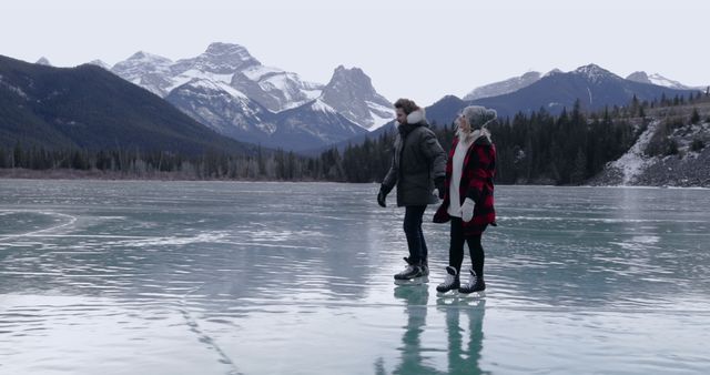 Couple enjoys ice skating on a frozen lake surrounded by picturesque mountains under overcast sky. Ideal for travel brochures, winter sport advertisements, romantic holiday promotions, and nature magazines showcasing adventure and scenic beauty.