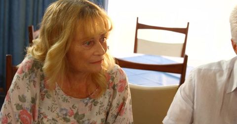 Senior woman in floral blouse looking thoughtful sitting at cozy backlit dining table
