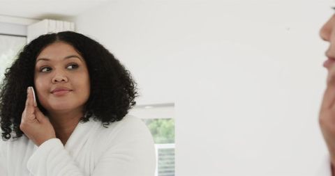 African American Woman Performing Morning Skincare Routine in White Bathrobe by Mirror