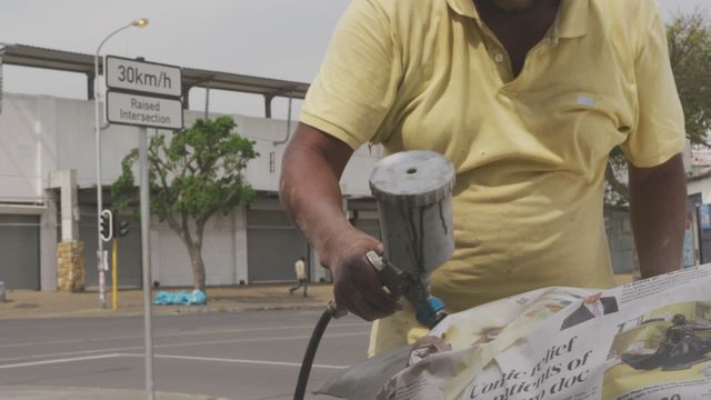 Close-up of a male panel beater using a spray gun for car painting, set in an urban landscape. Perfect for illustrating automotive repair processes, highlighting craftsmanship in car refurbishing. Utilized in études on urban artisans, mechanistic vocations, or features on repair industry specifics.