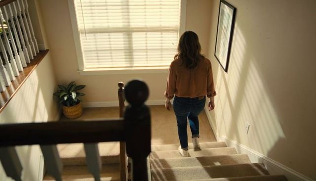 Woman descending sunlit carpeted staircase wearing jeans and sneakers