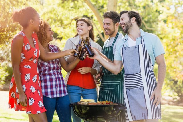Group of friends toasting a beer bottle while preparing barbecue grill in park