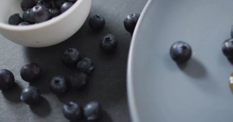 Glistening blueberries on dark matte surface with white bowl and gray ceramic plate