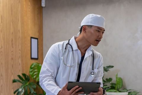 Asian male doctor wearing white coat scrub cap holding tablet and stethoscope in clinic