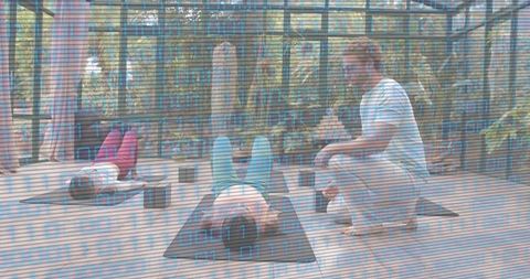 Yoga instructor guiding students through floor poses in glass studio with lush greenery
