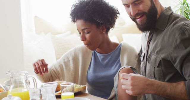 Interracial couple sitting at a breakfast table, engaging in intimate conversation while eating. This photograph can be used to reflect themes of love, diversity, and domestic life. Ideal for websites, advertisements, and articles focusing on relationships, morning routines, and inclusive living.