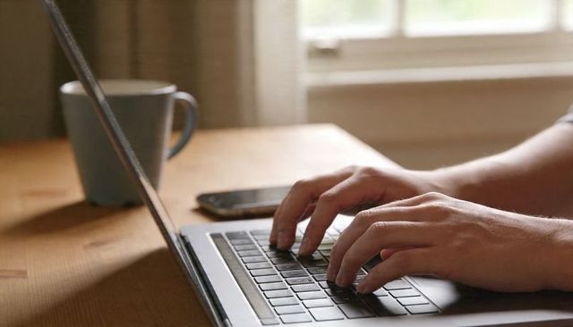 Typing adult hands on laptop at home with coffee mug and smartphone on wooden table