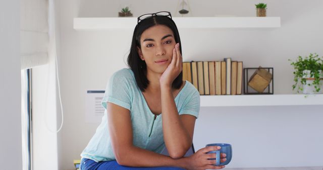 Young woman holding a coffee cup and sitting in a modern, minimalist living room with shelves in the background. Useful for lifestyle blogs, articles on mental health or wellness, advertisements for home decor products, or ads targeting young adults.