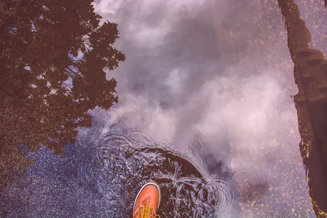 Brown boot stepping into water puddle showing reflections of trees and cloudy sky. Captures rainy weather, outdoor activities, and relaxed strolls. Suggests concepts of exploration, nature, climate, and reflection. Useful for lifestyle, adventure, travel, and weather-related content.
