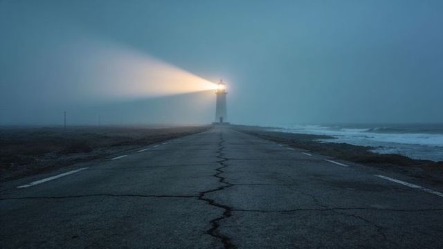 Lighthouse beacon piercing coastal fog over cracked road at twilight
