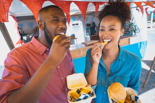 Happy diverse couple enjoying takeaway food, standing beside a food truck. They are smiling and appear to be having a good time. This image can be used for promoting street food events, food truck businesses, casual dining, and lifestyle blogs focusing on urban culture and social activities.