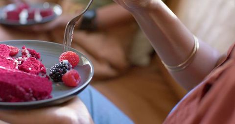 Woman enjoying raspberry cake on couch, forking bright magenta dessert with berries