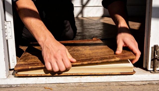 Kneeling hands revealing hidden ledger under wooden plank at sunlit doorway