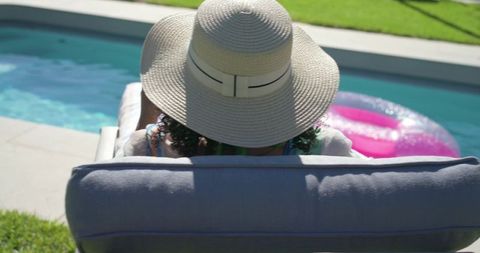 Woman relaxing and enjoying poolside lounge wearing wide brim straw hat near pink float
