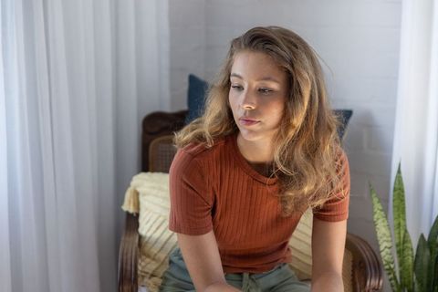 Thoughtful young woman sitting in rattan chair in sunlit minimalist living room interior