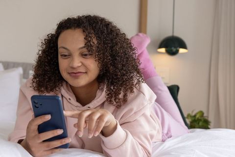 African american woman lying on bed tapping smartphone, cozy loungewear and ambient lamp