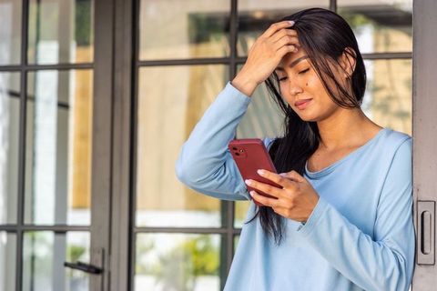 Asian woman standing in doorway using smartphone while touching hair, contemplative moment
