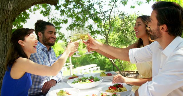 Friends sitting around a table with salads and toasting wine glasses during a daytime garden party. The scene is set under a large tree with lush greenery, evoking feelings of happiness, health, and social togetherness. Perfect for advertising social gatherings, summer events, and lifestyle content promoting friendship and celebration.