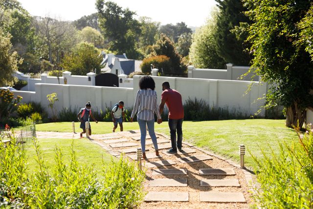 This image shows an African-American couple standing on a garden pathway, waiting to greet their children who are returning from school. The son and daughter are walking towards their house, creating a warm and welcoming scene. This image can be used for family-oriented content, educational materials, parenting blogs, and advertisements promoting family values and home life.