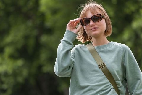 Woman adjusting tortoiseshell sunglasses in park wearing sage sweatshirt and olive strap