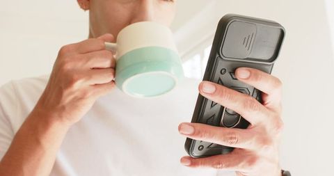 Man sipping coffee while checking smartphone near sunlit window with mint green mug