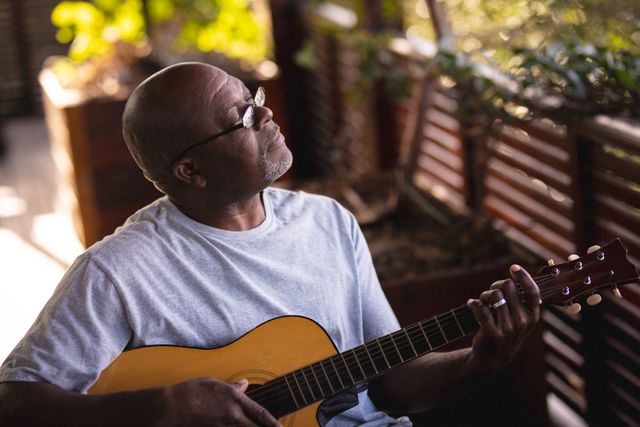 Relaxed african american senior man sitting in patio and playing guitar. retirement lifestyle, spending time alone at home.