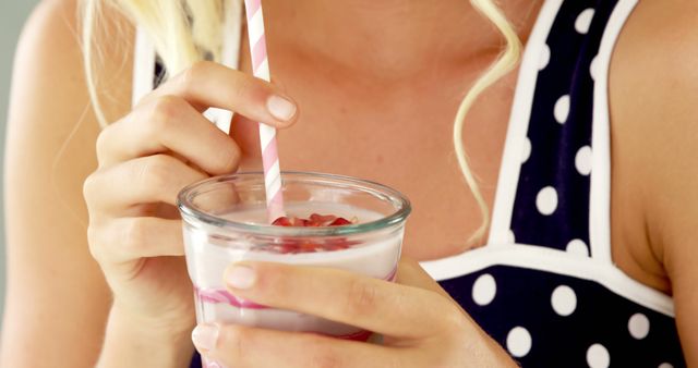 Young blonde woman holding yogurt smoothie with a striped straw. Close-up on hands and drink. Visible blonde hair curls and polka dot strap top. Great for health, food and beverage promotions, summer refreshments advertisement, and lifestyle blogs.