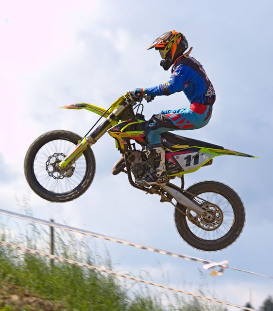Motocross rider performing jump during race on dirt track with blue sky in background. Scene shows action, adrenaline, and extreme sportsmanship. Useful for illustrating extreme sports, adventure tourism, motorbike events, lifestyle magazines, and action-themed advertisements.