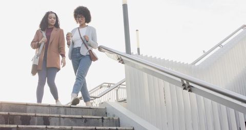 Two women walking down city stairs holding coffee and carrying shoulder bags