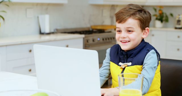 Caucasian boy smiles while using a laptop at home. He's engaged in an online learning session in a bright kitchen setting.