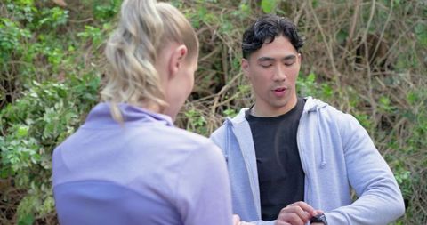 Asian man checking smartwatch while female companion listening on woodland fitness trail