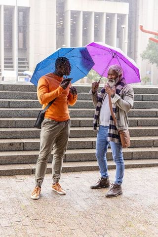 Urban colleagues standing in rain holding colorful umbrellas while checking phone