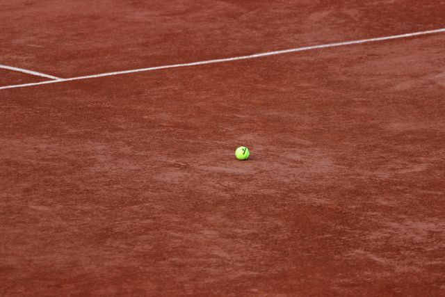 Tennis ball lying still on red clay court, with clear white boundary lines. Suitable for content related to tennis, sports promotions, training guides, and inspirational sports concept. Perfect for illustrating calm moments between game plays or highlighting the texture and composition of a clay tennis court.