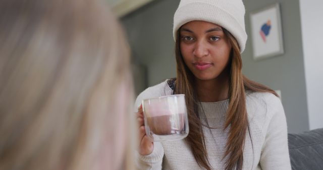 Young woman wearing a beanie and sweater, enjoying a hot drink in a cozy home environment. Perfect for use in lifestyle blogs, home comfort advertisements, social media posts about relaxation or cozy living, and marketing materials for beverages.