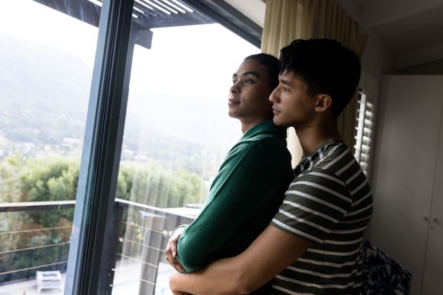 Romantic multiracial gay couple embracing and looking through window at home, copy space. Contemplation, affectionate, unaltered, love, togetherness, homosexual, lifestyle and home concept.