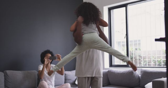 Family enjoying a joyful moment at home as father lifts daughter while mother captures the memory on phone. Ideal for promoting family bonding, happiness, modern parenting, and home lifestyle services.