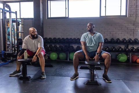 Two african american men sitting and laughing on gym benches, talking after workout