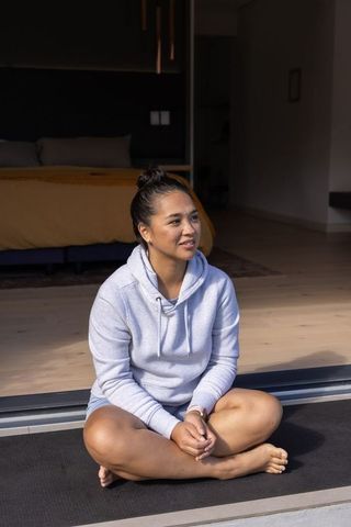 Mid adult asian woman sitting cross-legged in hoodie at sliding doorway for yoga and wellness