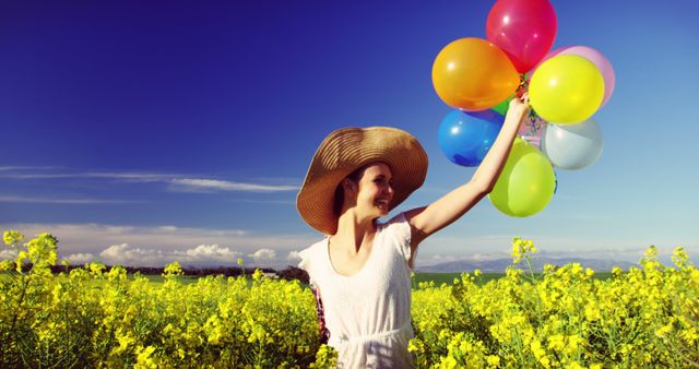 This image features a joyous woman in a wide-brimmed hat holding colorful balloons in a yellow flower field under a clear blue sky. Vibrant and cheerful, this picture makes a great choice for marketing materials promoting happiness, nature, outdoor activities, or spring events.