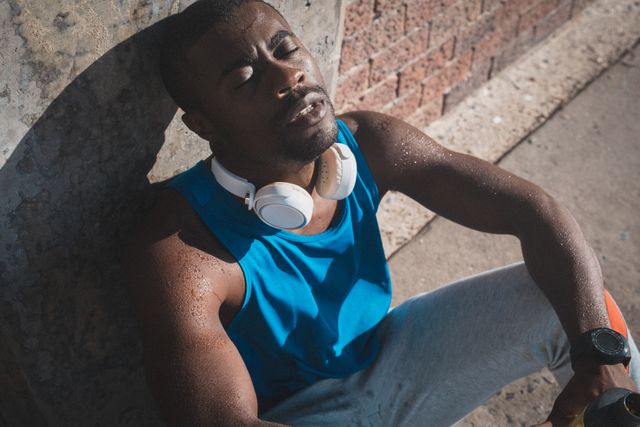 African American man sitting against wall, resting after intense workout. Wearing headphones and athletic clothing, sweating under the sun. Ideal for use in fitness, health, and lifestyle content, promoting exercise, recovery, and outdoor activities.