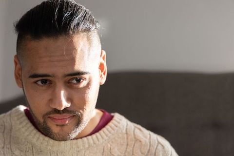 Indian mid-adult man sitting on sofa looking contemplative in cream knit sweater wearing top knot