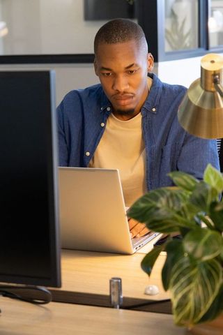 African american man working on laptop in modern coworking office with desk plant lamp
