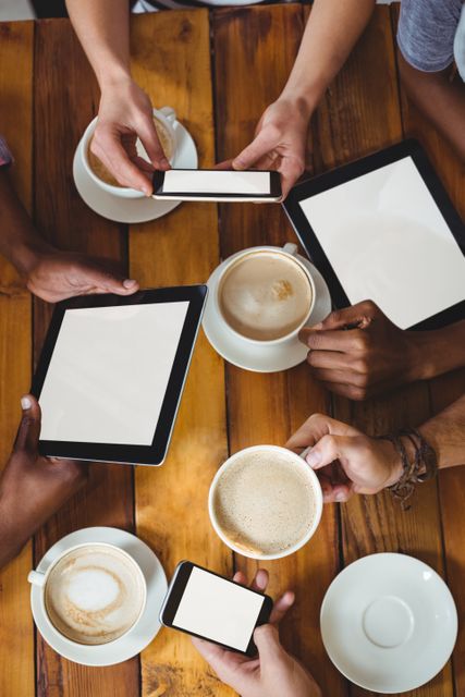 Group of friends sitting around wooden table, holding smartphones and tablets while enjoying coffee. They are engaging in a casual social gathering, showcasing modern communication and technology usage. Ideal for depictions of social media use, technology in daily life, or casual meetings in cafes.
