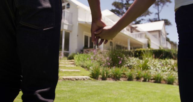 Rear midsection of diverse couple holding hands outside house in sunny garden, copy space. Summer, home, romance, relationship, togetherness, domestic life and lifestyle, unaltered.