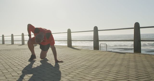 Man in fitness attire stretching on boardwalk by the ocean after jogging. Perfect for use in articles about outdoor exercise routines, fitness for retired individuals, seaside workout tips, or promotional materials for fitness apparel. Captures theme of maintaining health and wellness in senior years.